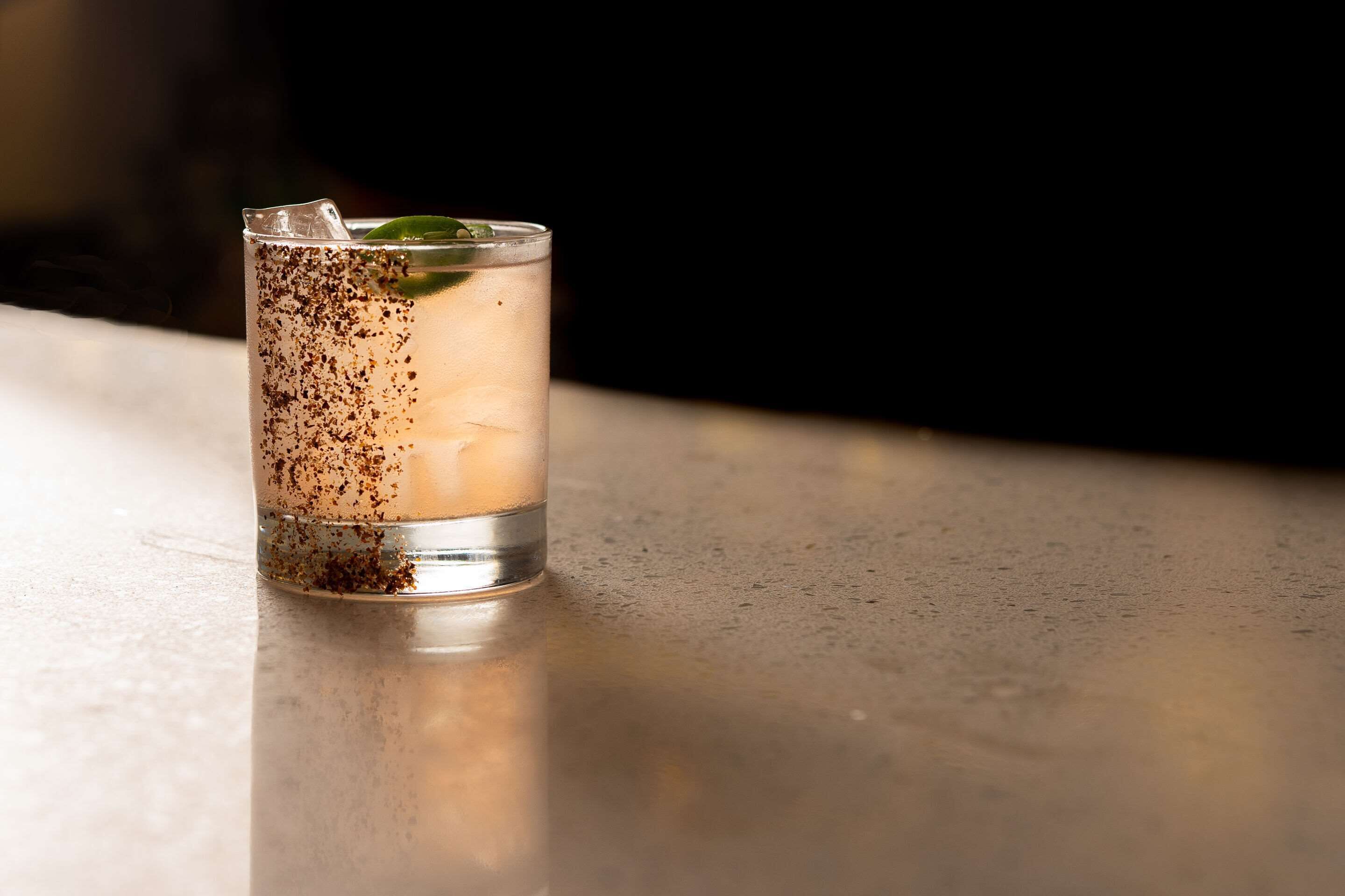 A clear glass with a beverage, ice cubes, and a mint leaf on a reflective surface.