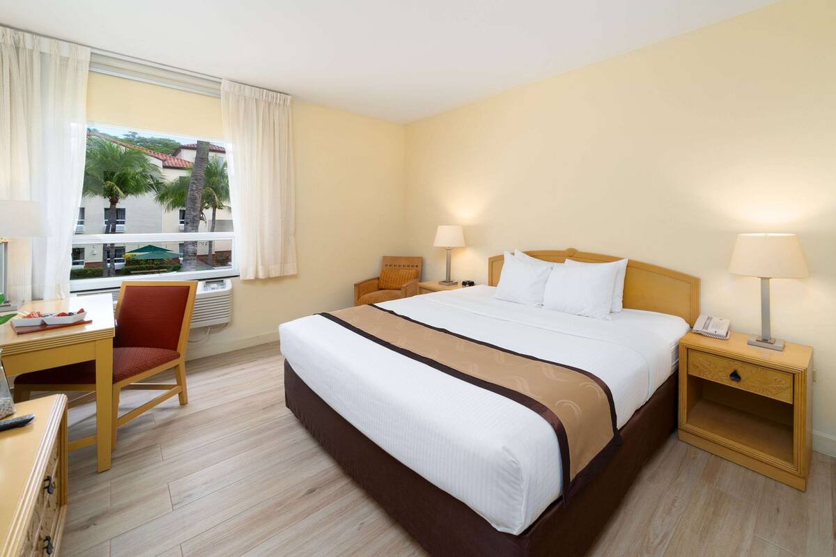 A hotel room with a large bed, a desk with a chair, a nightstand with a lamp, and a window with curtains.