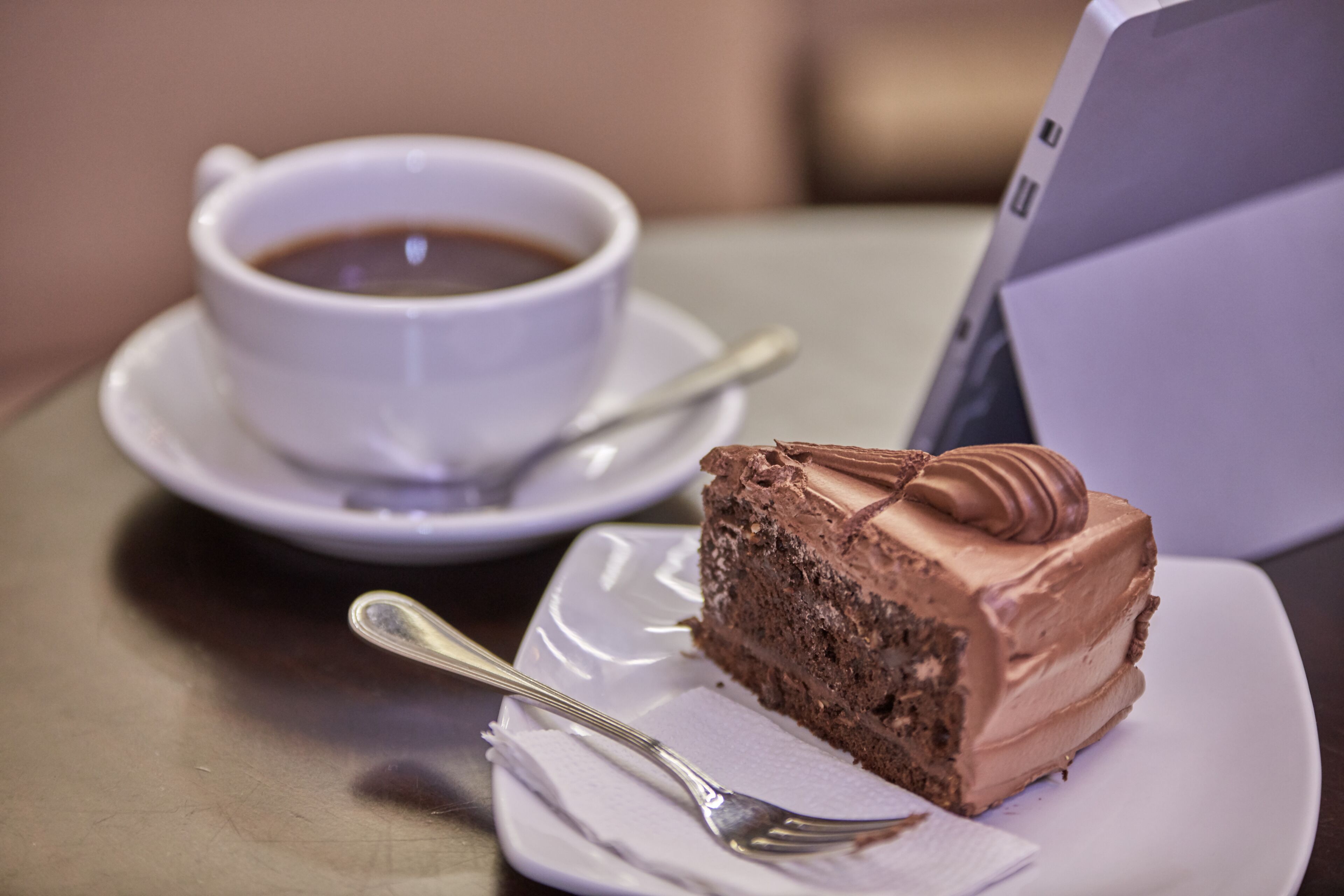 Una porción de pastel de chocolate sobre un plato blanco, con una cuchara y una servilleta, junto a una taza de café en un platito.