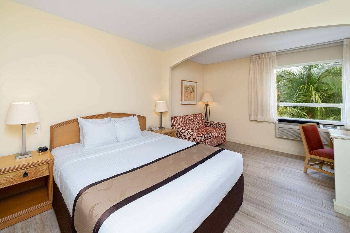 A hotel room with a large bed, two bedside tables, a desk, a chair, and a window with a view of palm trees.