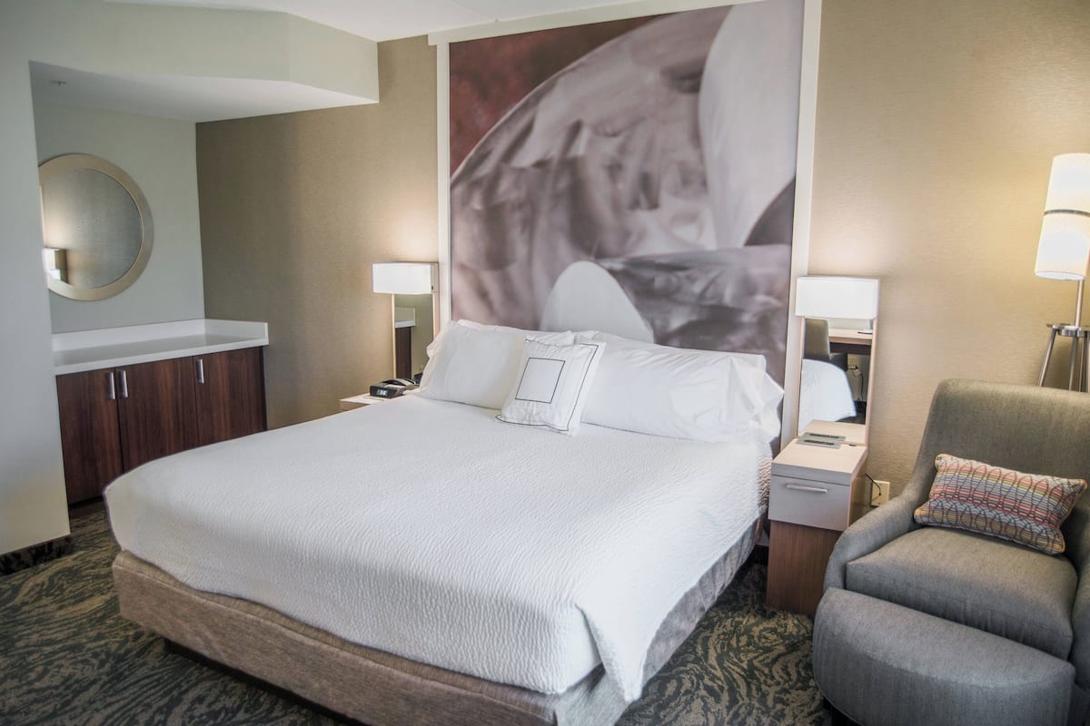 A hotel room with a large bed, a desk with a lamp, a chair, a wardrobe, and a wall-mounted mirror.