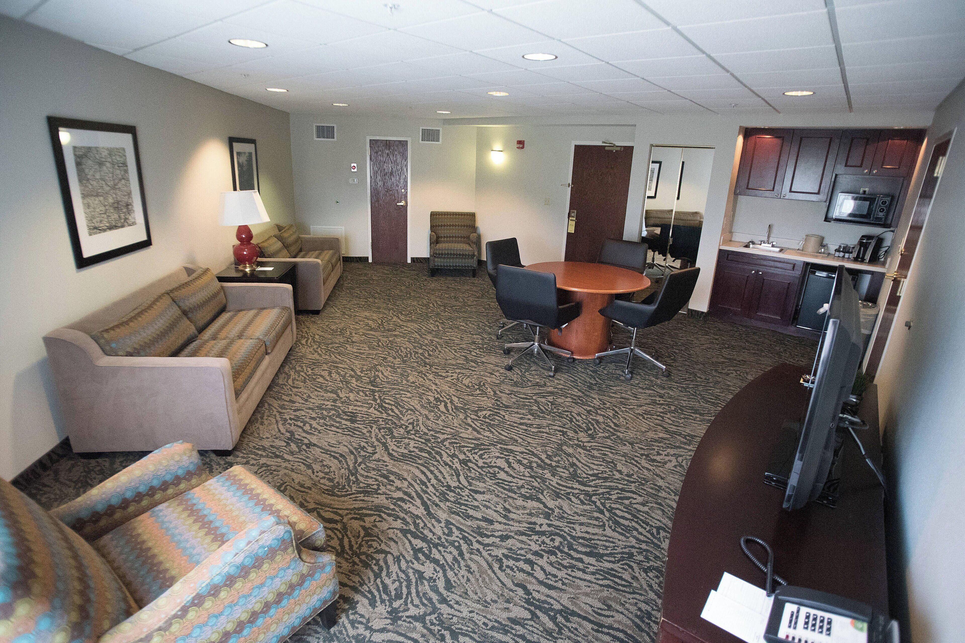 A hotel room with a sofa, armchair, dining table, chairs, a small kitchenette, and a television.