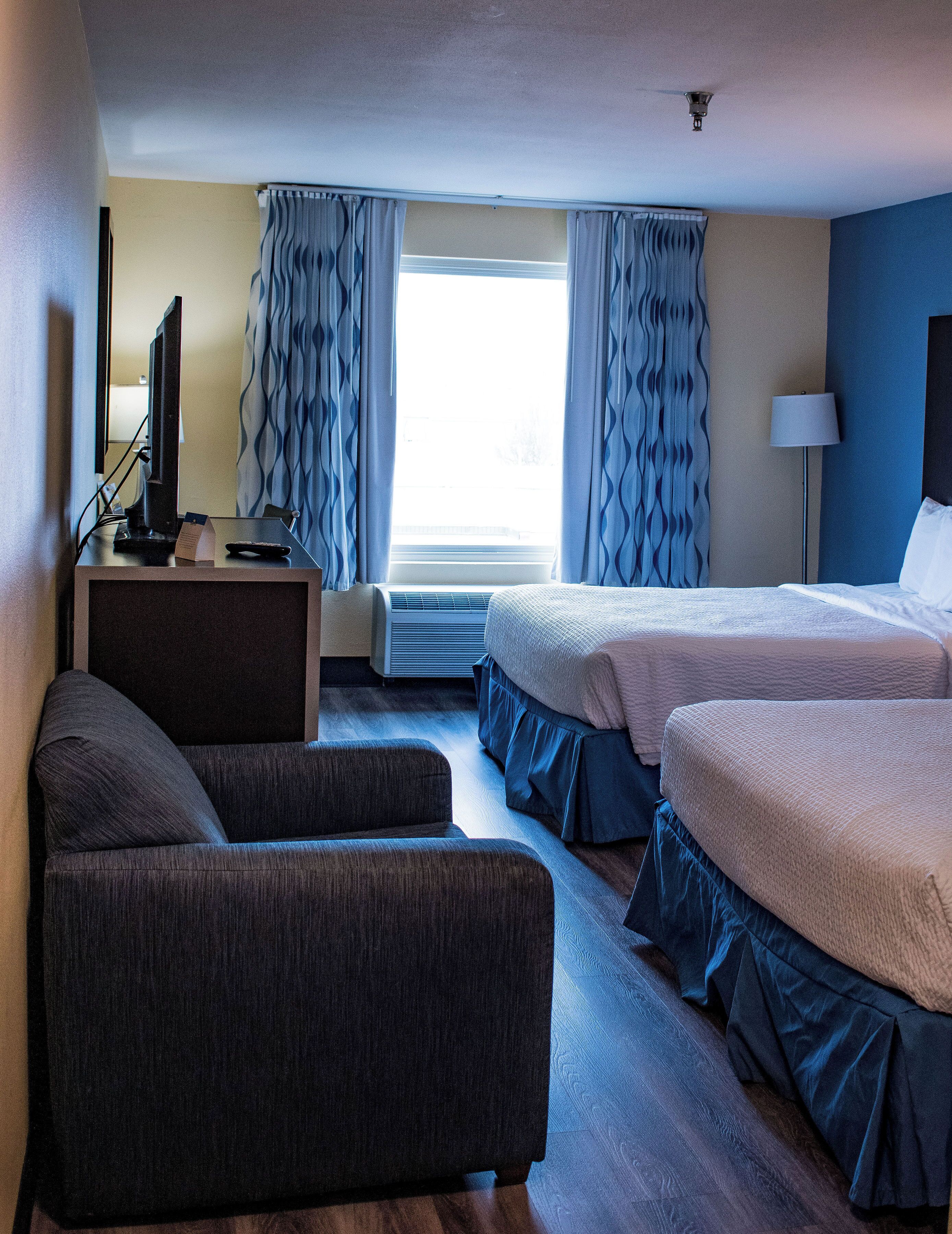 Una habitación de hotel con escritorio, dos camas, una silla, lámpara y ventana con cortinas.