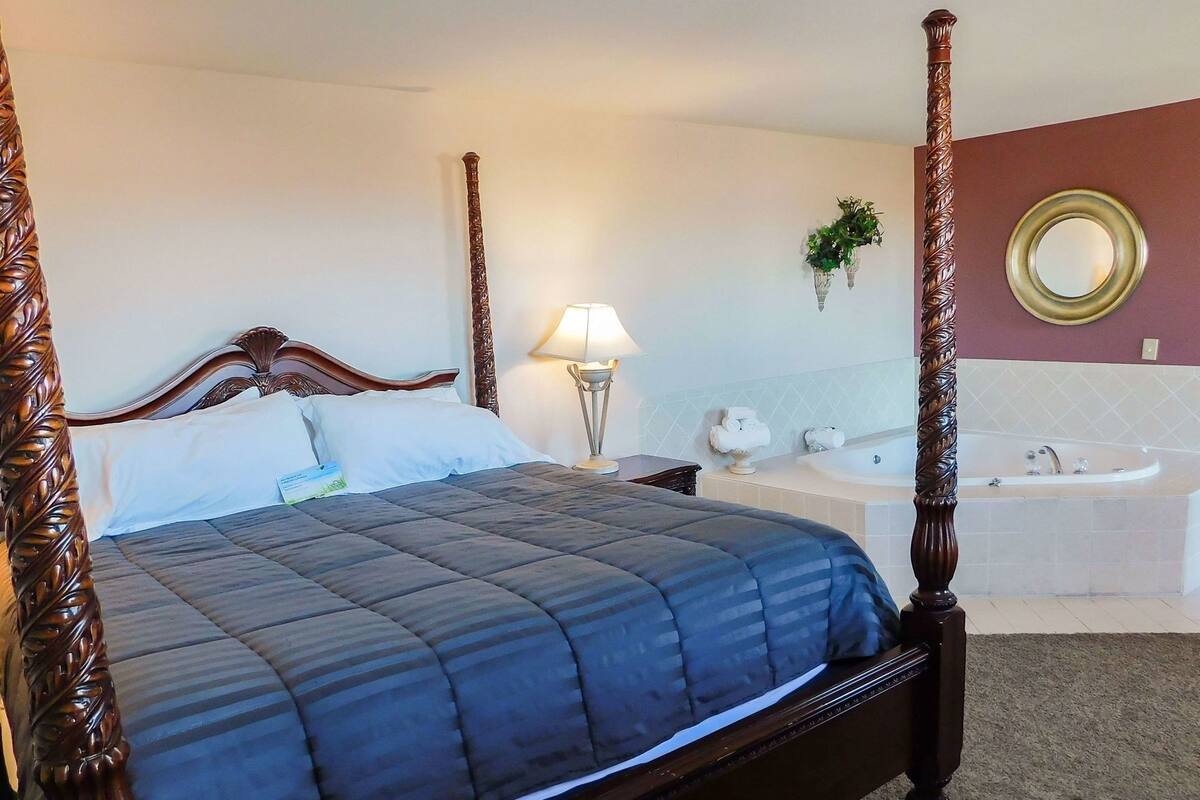 A four-poster bed with a dark blue quilt, a bedside lamp, a white bathtub, and a round mirror on the wall.