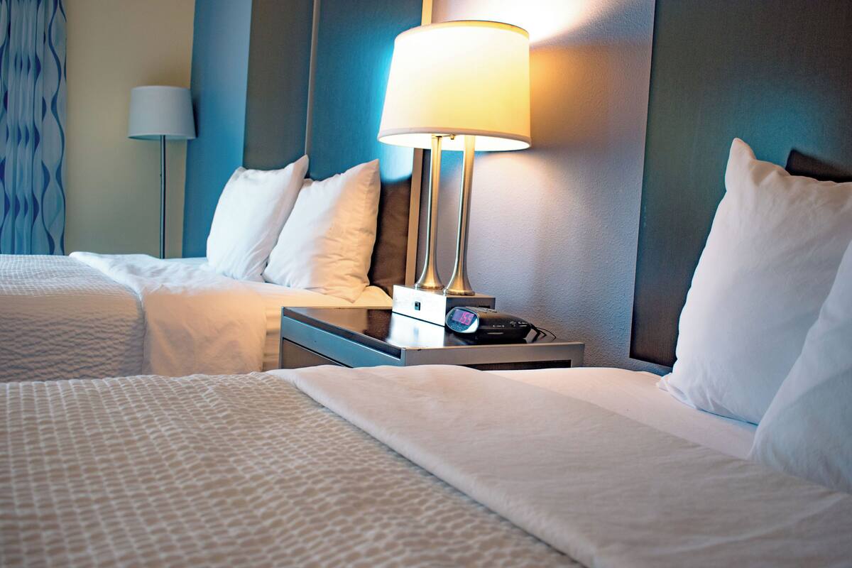 A hotel room with two beds, a bedside table with a lamp, and a digital clock.