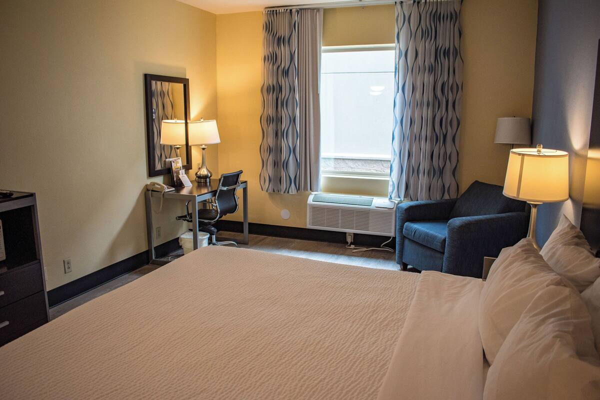 A hotel room with a large bed, a desk with a telephone, two chairs, a window with curtains, and two lamps.