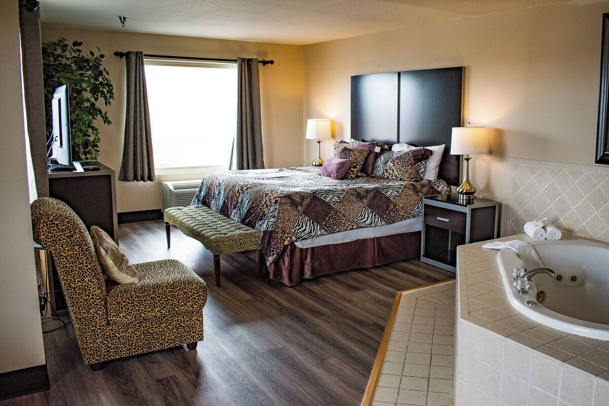 A hotel room with a bed, a sitting area with a chair, a desk, a lamp, a large mirror, and a bathtub.