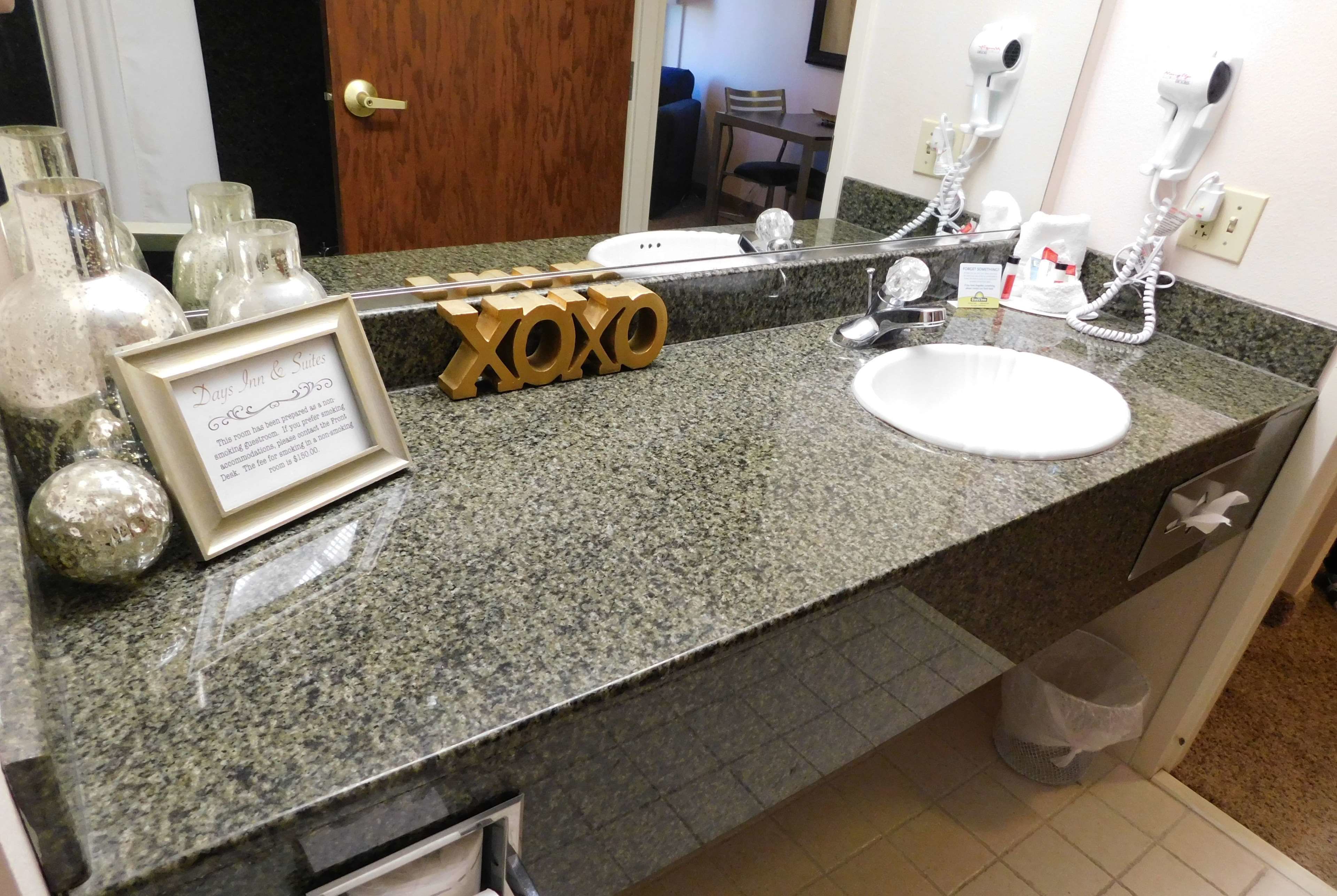 A bathroom with a granite countertop, a sink, and a mirror. There are hair dryers mounted on the wall, a framed sign, and a trash can.