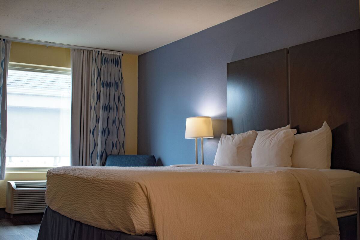 A hotel room with a bed, a lamp, a window with curtains, and an air conditioning unit.