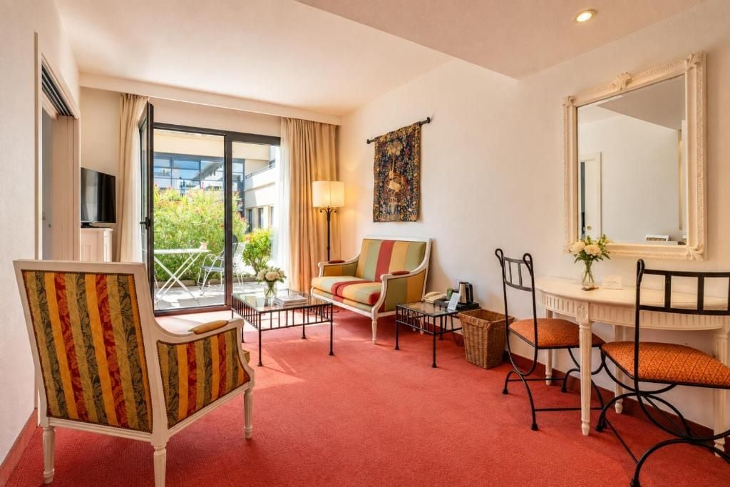A hotel room with a sofa, armchair, dining table, chairs, a mirror, and a television.