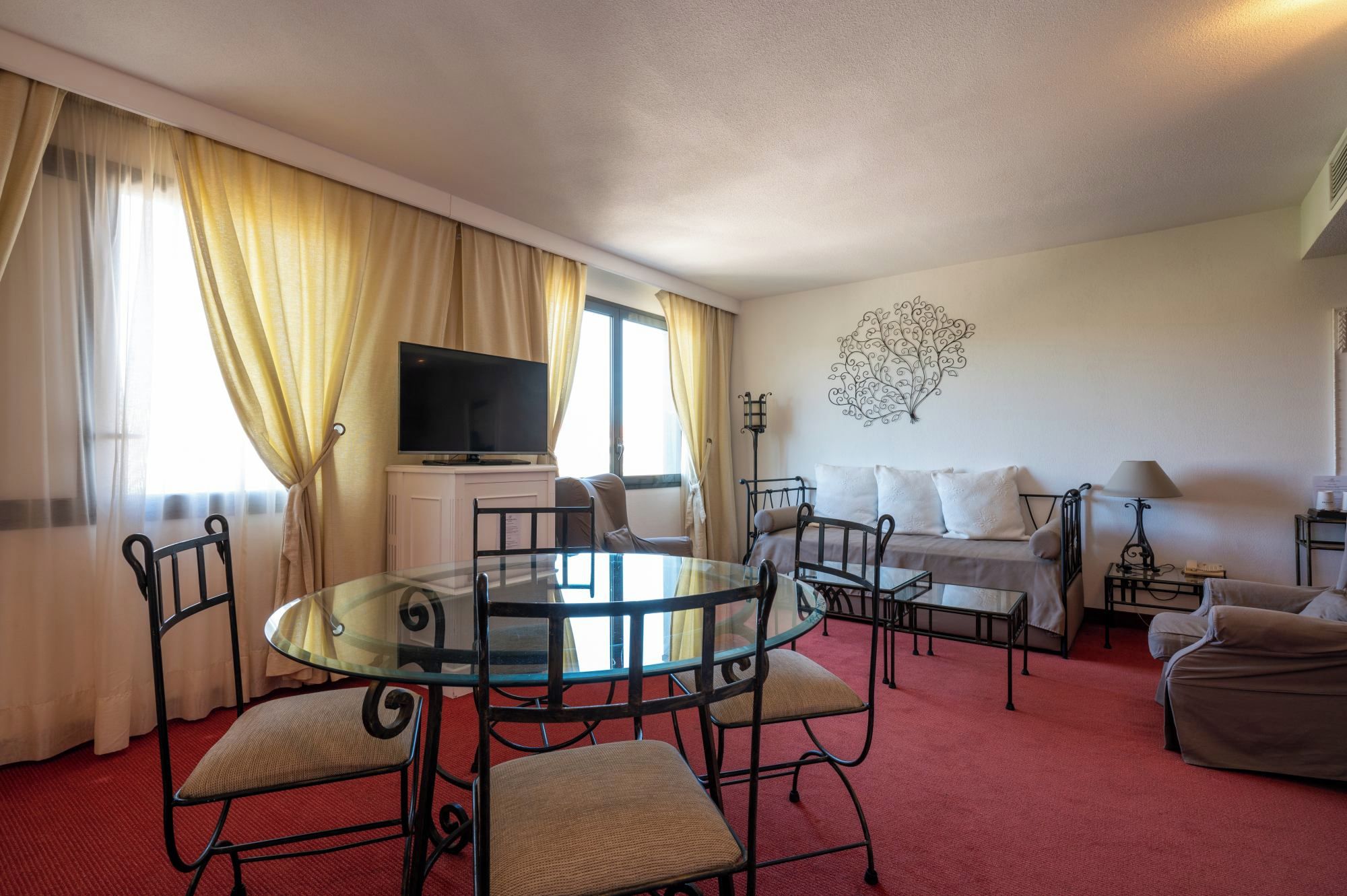 A hotel room with a glass dining table, a bed with white linens, a television on a cabinet, and a decorative wall piece.