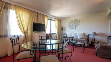 A hotel room with a glass dining table, a bed with white linens, a television on a cabinet, and a decorative wall piece.