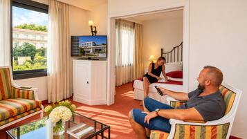 A hotel room with a sofa, armchair, a small table with a vase and book, a TV on the wall, and a view of a building outside.