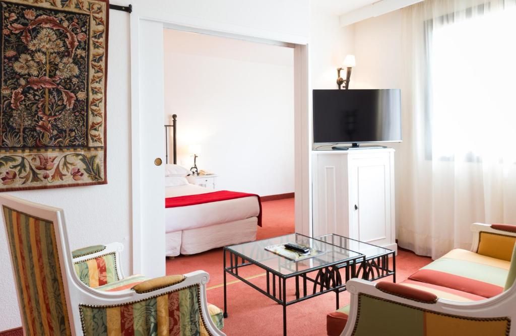 A hotel room with a bed, a television, a glass coffee table, and a seating area with a sofa and armchairs.