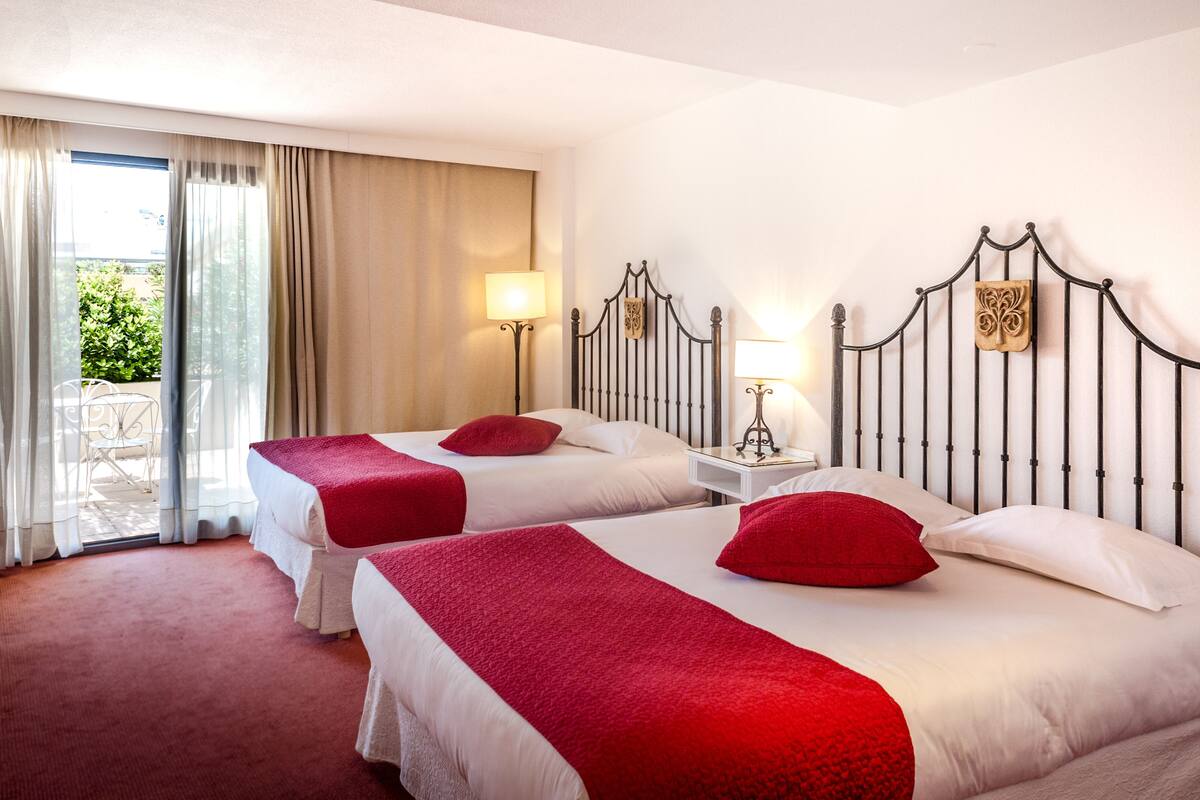 Ett hotellrum med två sängar, var och en med ett rött täcke och en kudde, ett nattduksbord med en lampa och utsikt över en balkong med ett bord och stolar.