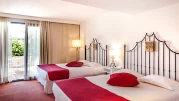 A hotel room with two beds, each with a red blanket and pillow, a nightstand with a lamp, and a view of a balcony with a table and chairs.