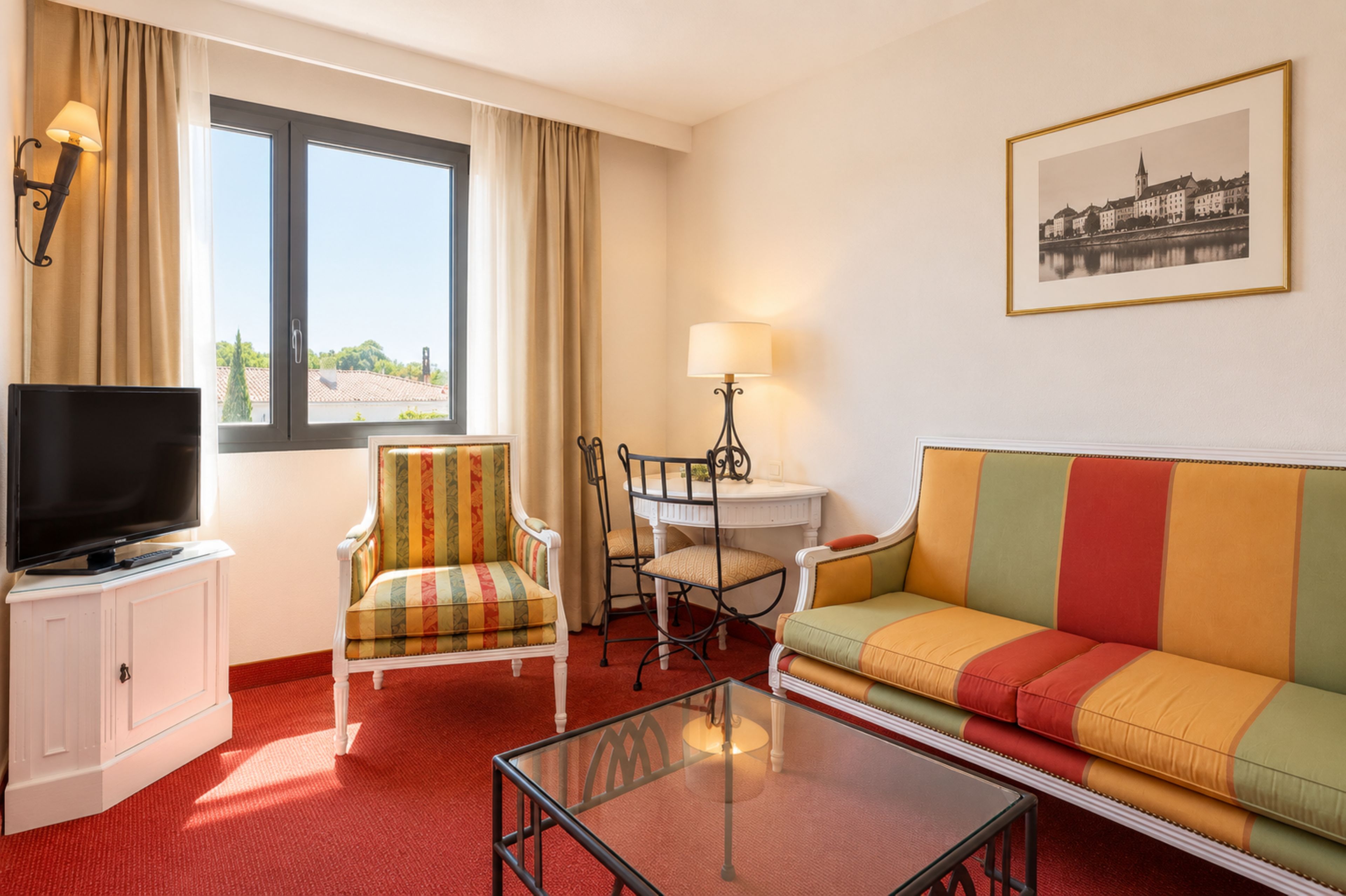 Una habitación de hotel con televisión, un sofá, una silla, una mesita y una ventana con cortinas.