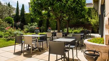 Une terrasse extérieure avec des tables et des chaises, entourée de verdure et agrémentée d’un grand vase rempli de fleurs.