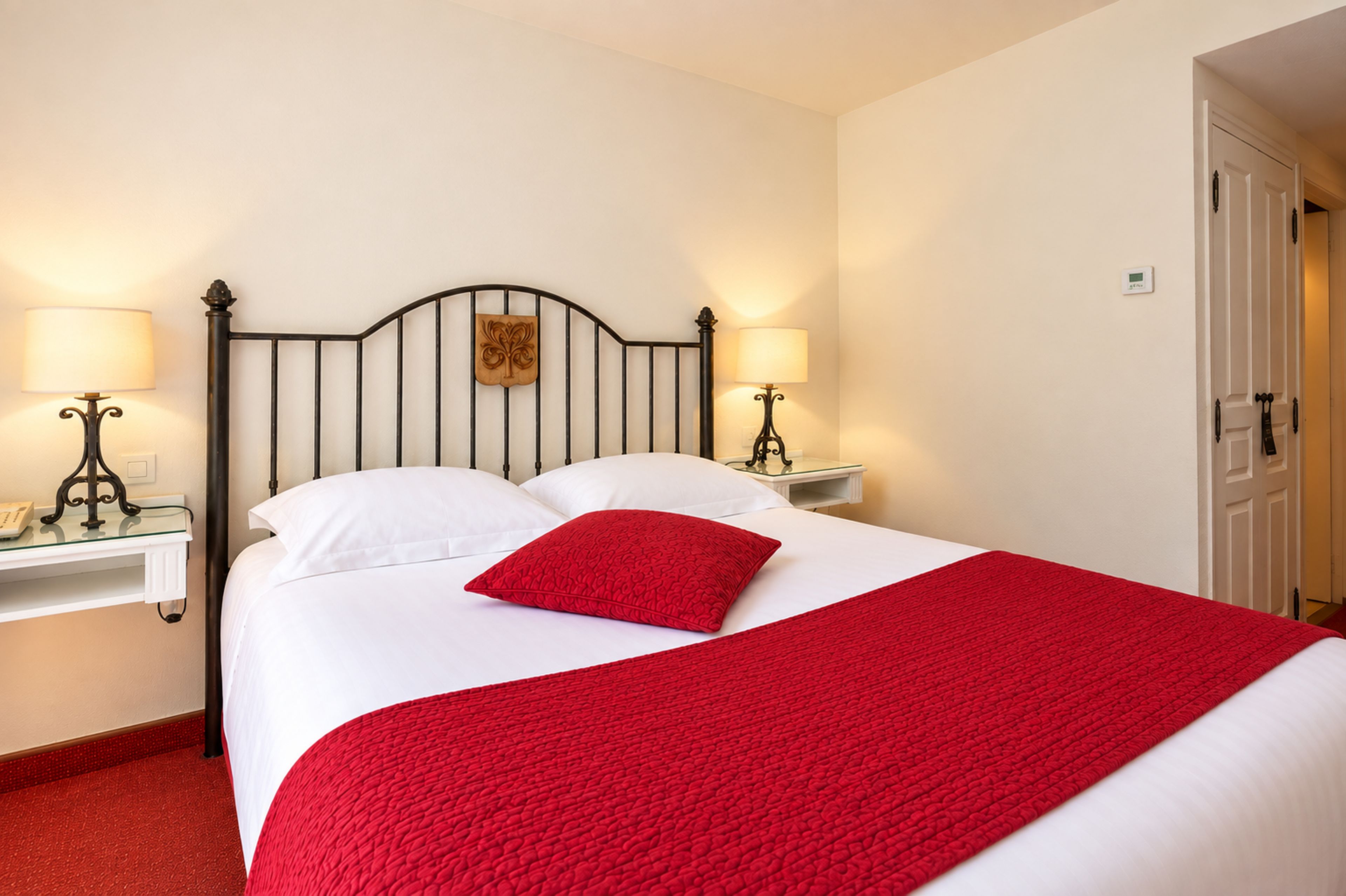 A bed with white bedding and a red throw, flanked by two bedside tables with lamps, in a room with a white door and walls.