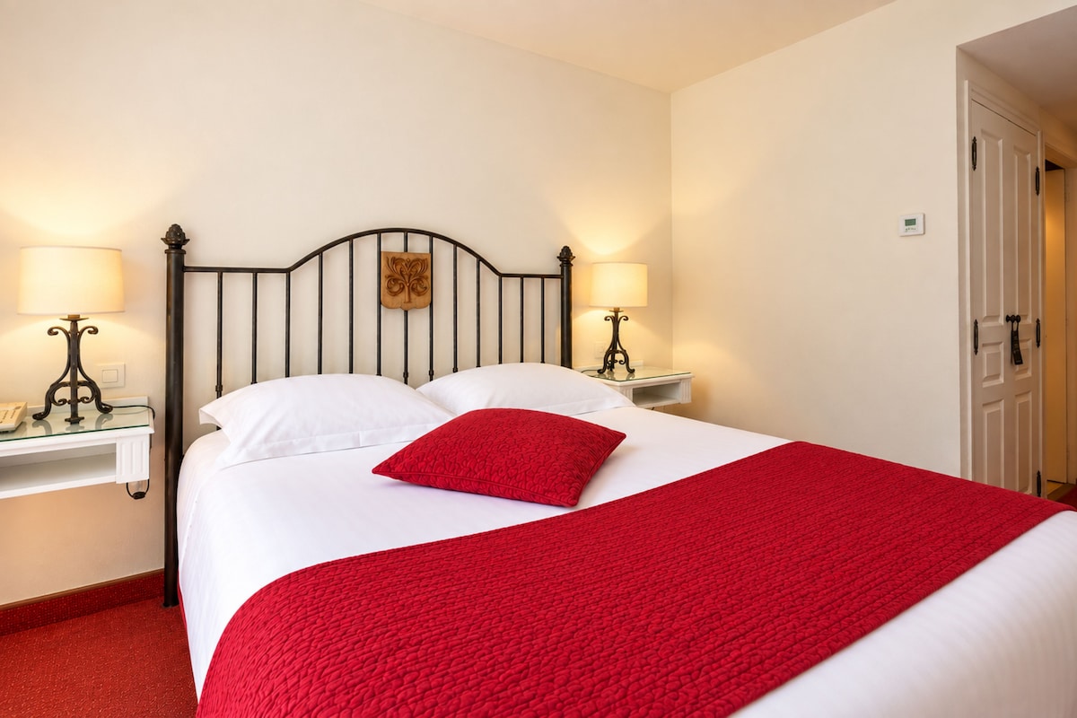 A bed with white bedding and a red throw, flanked by two bedside tables with lamps, in a room with a white door and walls.