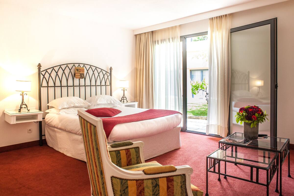Ett hotellrum med en säng, två sängbord med lampor, en stol, ett litet bord med en vas med blommor och en skjutdörr i glas som leder ut till en balkong.