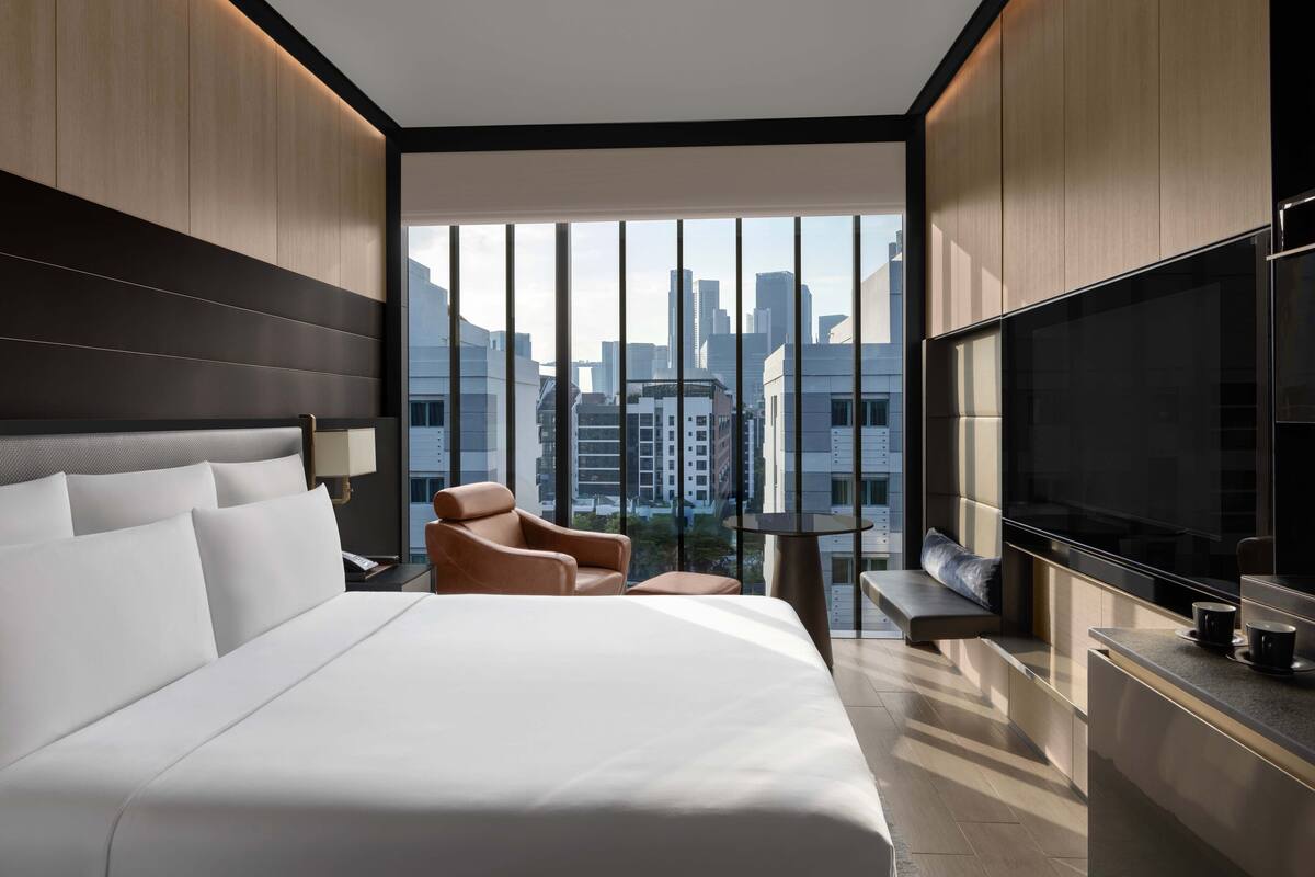 Una habitación de hotel moderna con una cama grande, una zona de estar con una silla y un banco, un televisor y vistas a la ciudad a través de amplias ventanas.
