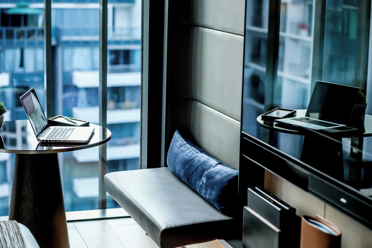 Una habitación de hotel moderna con un ventanal grande, un sofá, una mesita con una computadora portátil y otra mesita con una computadora portátil.