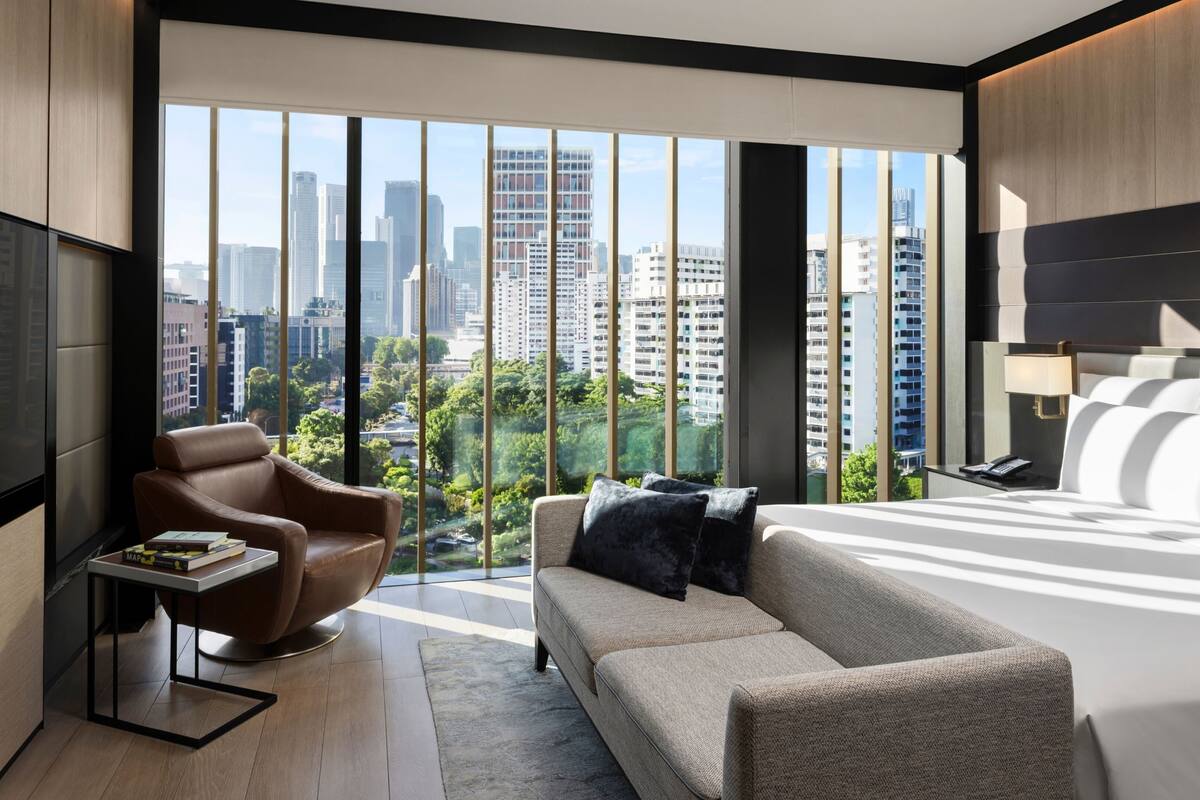 Una habitación de hotel moderna con un ventanal amplio que ofrece vistas a la ciudad, un sofá, una silla y un televisor.