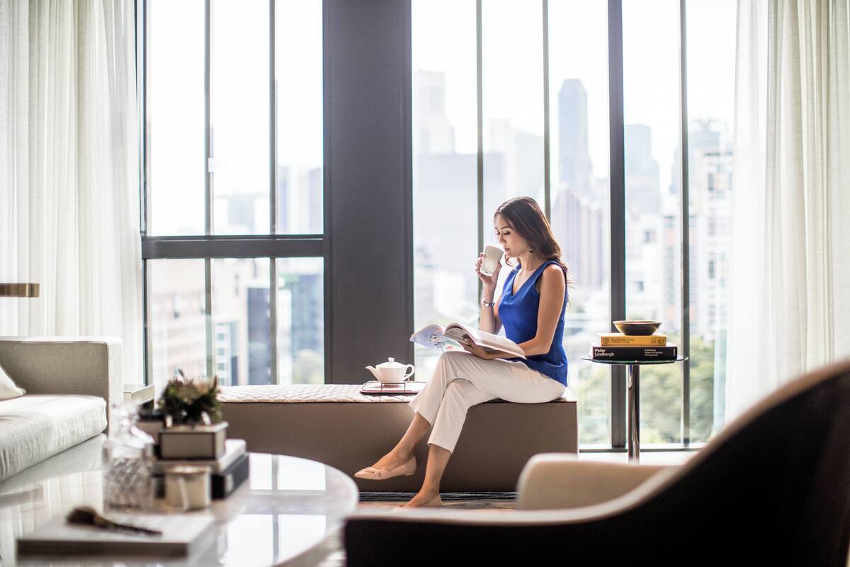 Una mujer sentada en un sofá junto a una ventana con vista a la ciudad, leyendo un libro.