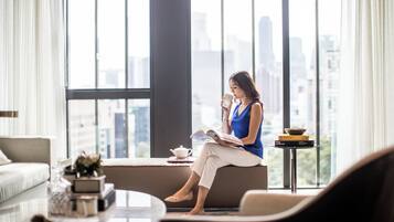 Una mujer sentada en un sofá junto a una ventana con vista a la ciudad, leyendo un libro.