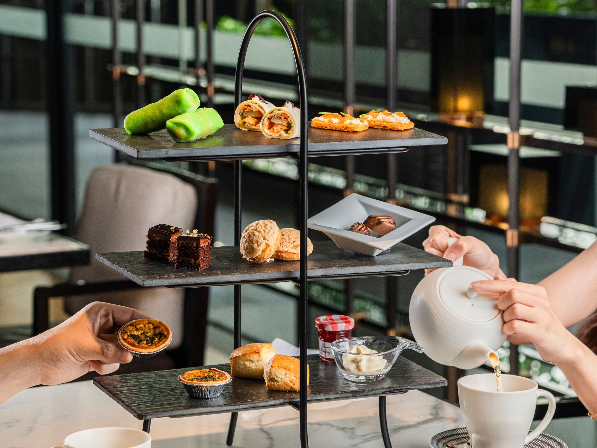 Un plateau à étages garni de diverses pâtisseries et desserts, une personne versant du thé dans une tasse, et un petit bol de beurre sur une table.