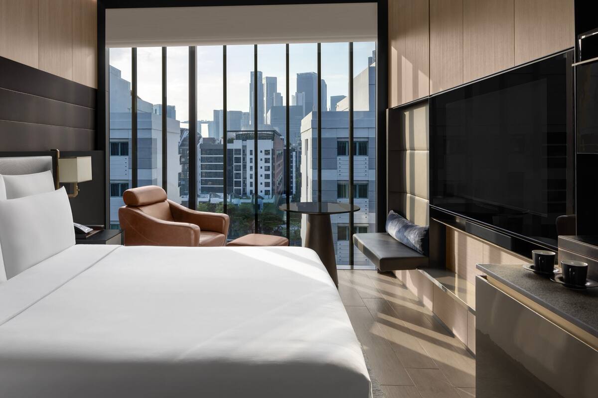 Una habitación de hotel moderna con una cama grande, un sillón de cuero, un televisor de pantalla plana y vistas a la ciudad a través de ventanales de gran altura.