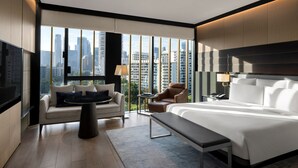Een moderne hotelkamer met een groot bed, een bank, een klein tafeltje en een stoel. De kamer heeft grote ramen met uitzicht op de stad.