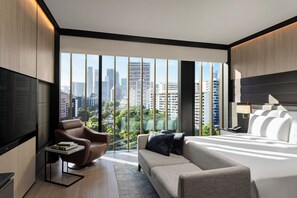Une chambre d’hôtel moderne, dotée d’un grand lit, d’un canapé, d’un fauteuil et d’une télévision. La chambre dispose de grandes fenêtres offrant une vue sur la ville.