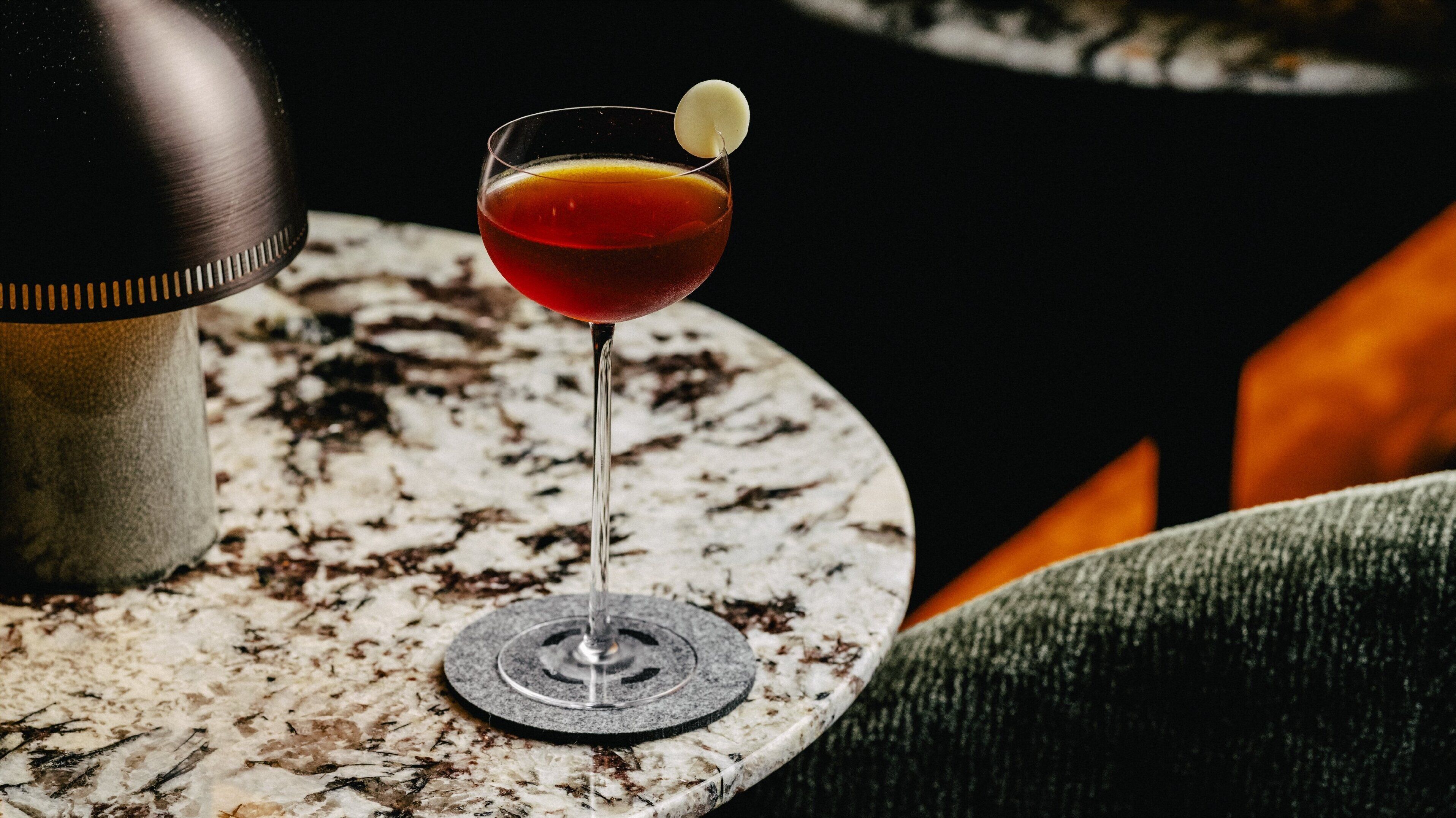 A martini glass with a lemon twist on a marble coaster, placed on a round table with a textured surface.