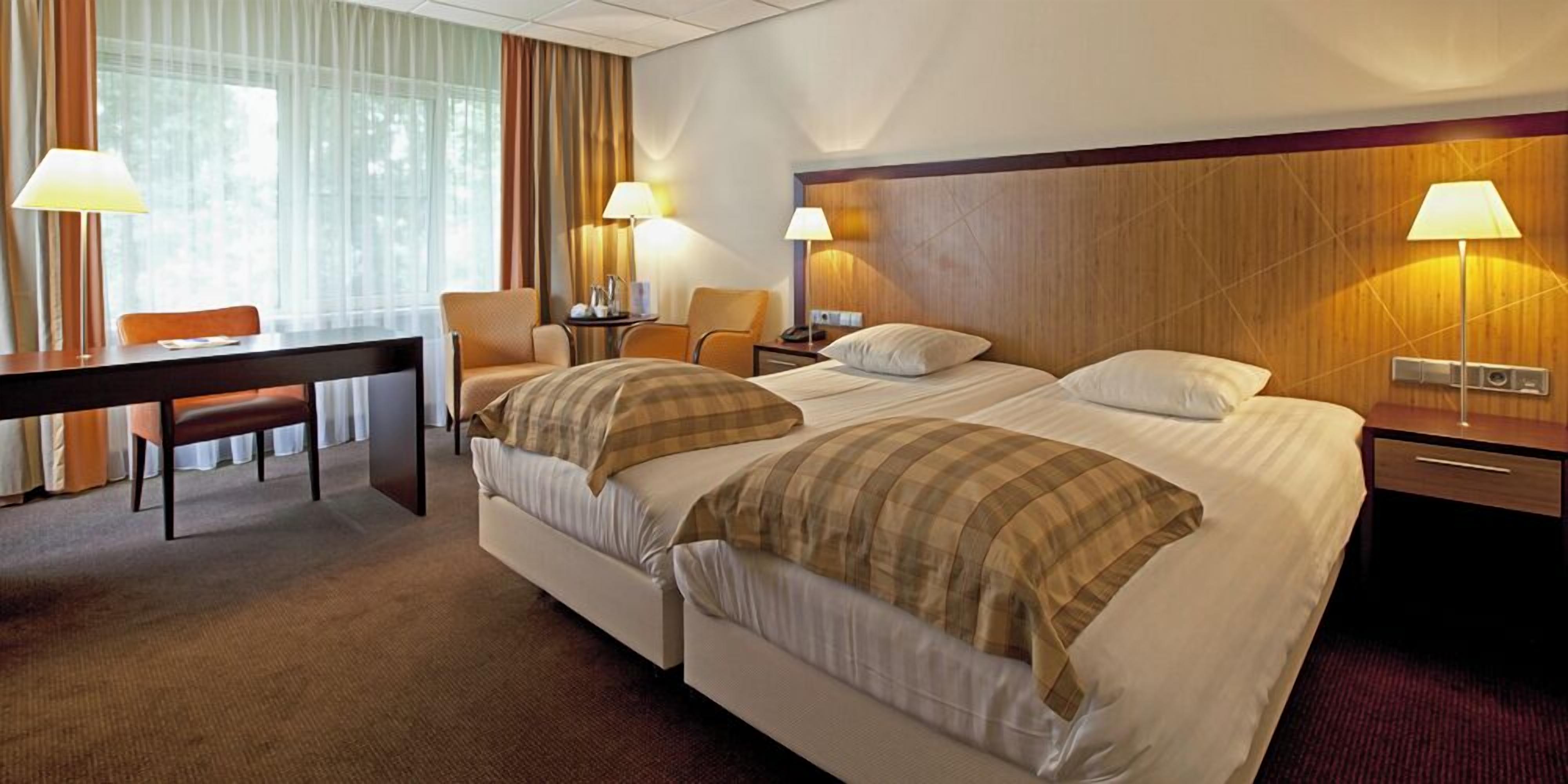 A hotel room with two beds, a desk, chairs, lamps, and a view of the outdoors.