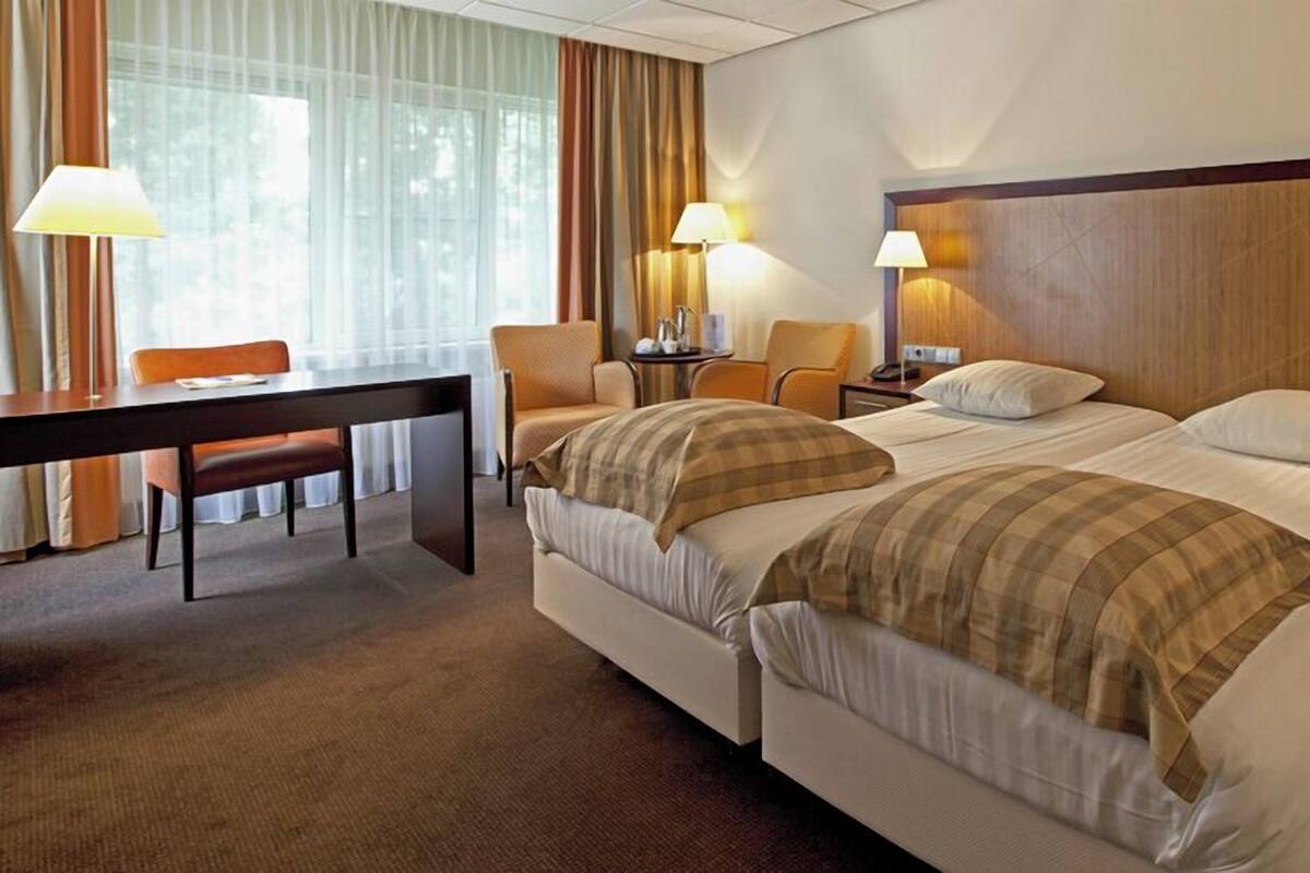 A hotel room with two beds, a desk, chairs, lamps, and a view of the outdoors.