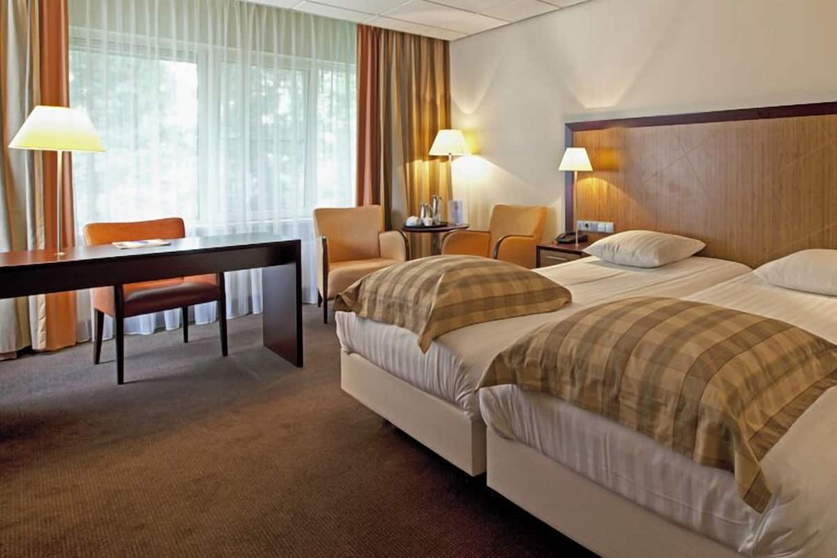 A hotel room with two beds, a desk, chairs, lamps, and a view of the outdoors.