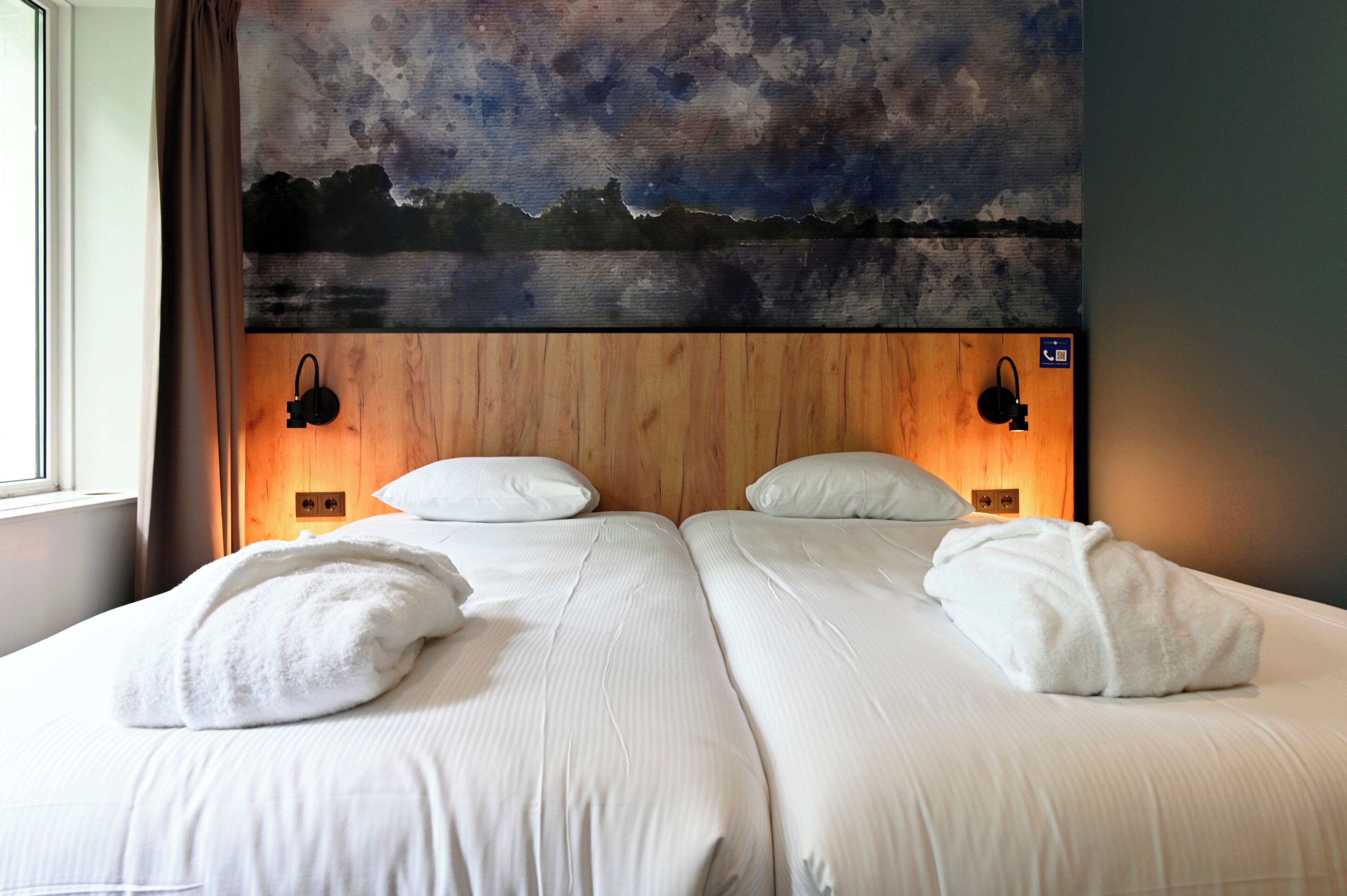 Cama doble con sábanas y almohadas blancas, cabecera de madera con un cuadro y dos lámparas fijadas a la pared.