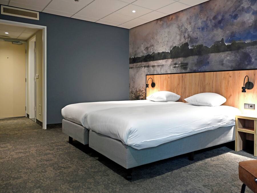 Una habitación de hotel con una cama grande, un cabecero con decoración en la pared, una mesita de noche y una puerta que da a otra habitación.