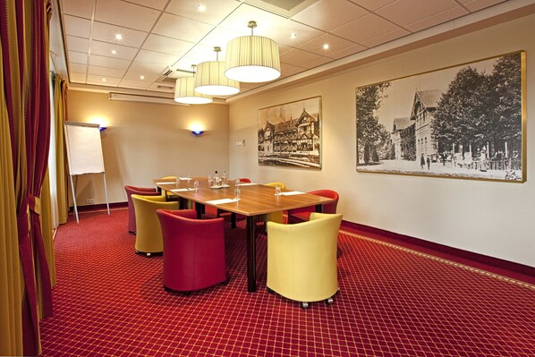 A conference room with a red carpet, a wooden table, and colorful chairs. There are two wall-mounted lamps and a framed black and white photo on the wall.