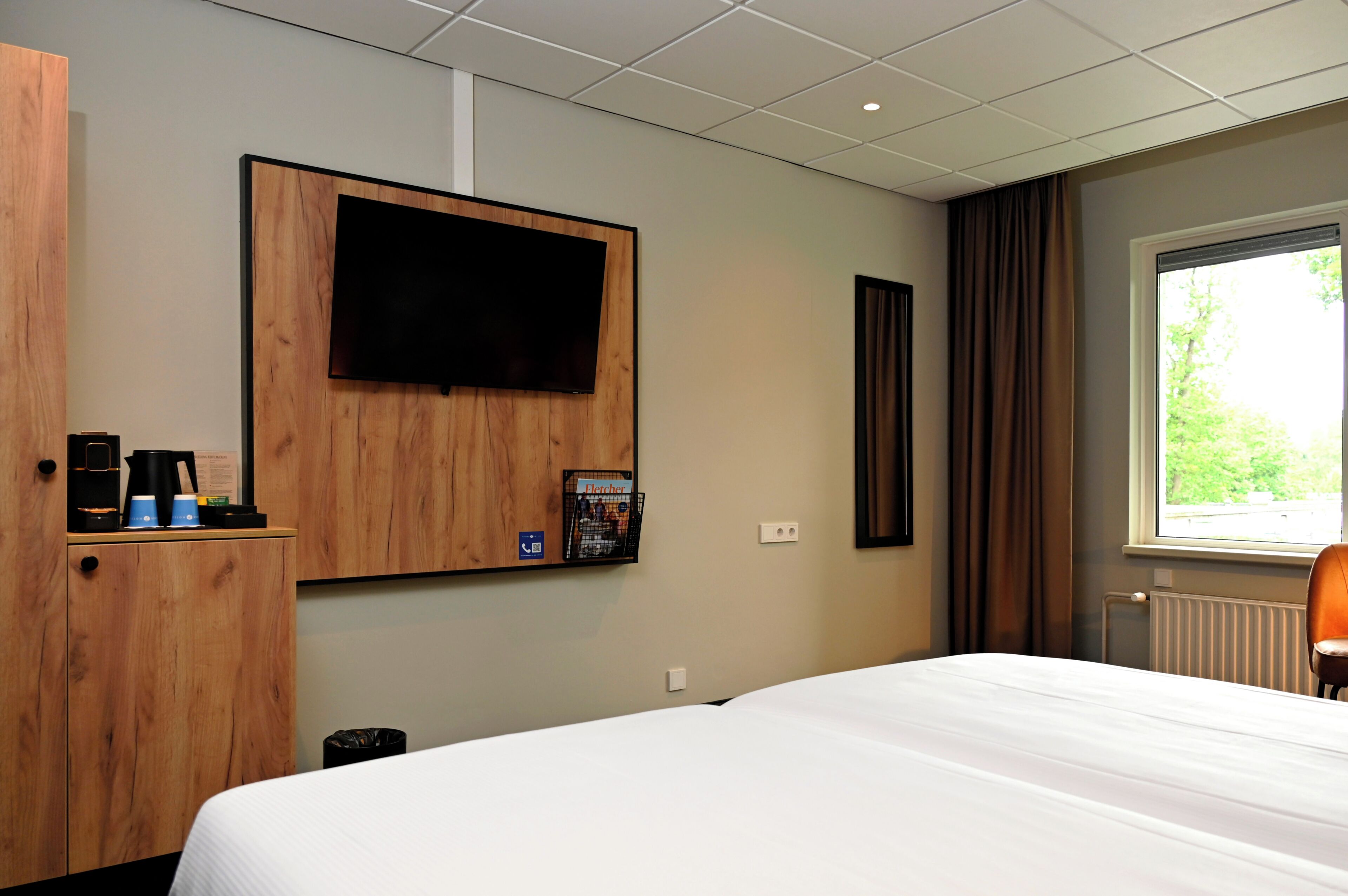 A hotel room with a bed, a mounted television, a coffee machine, and a window with curtains.