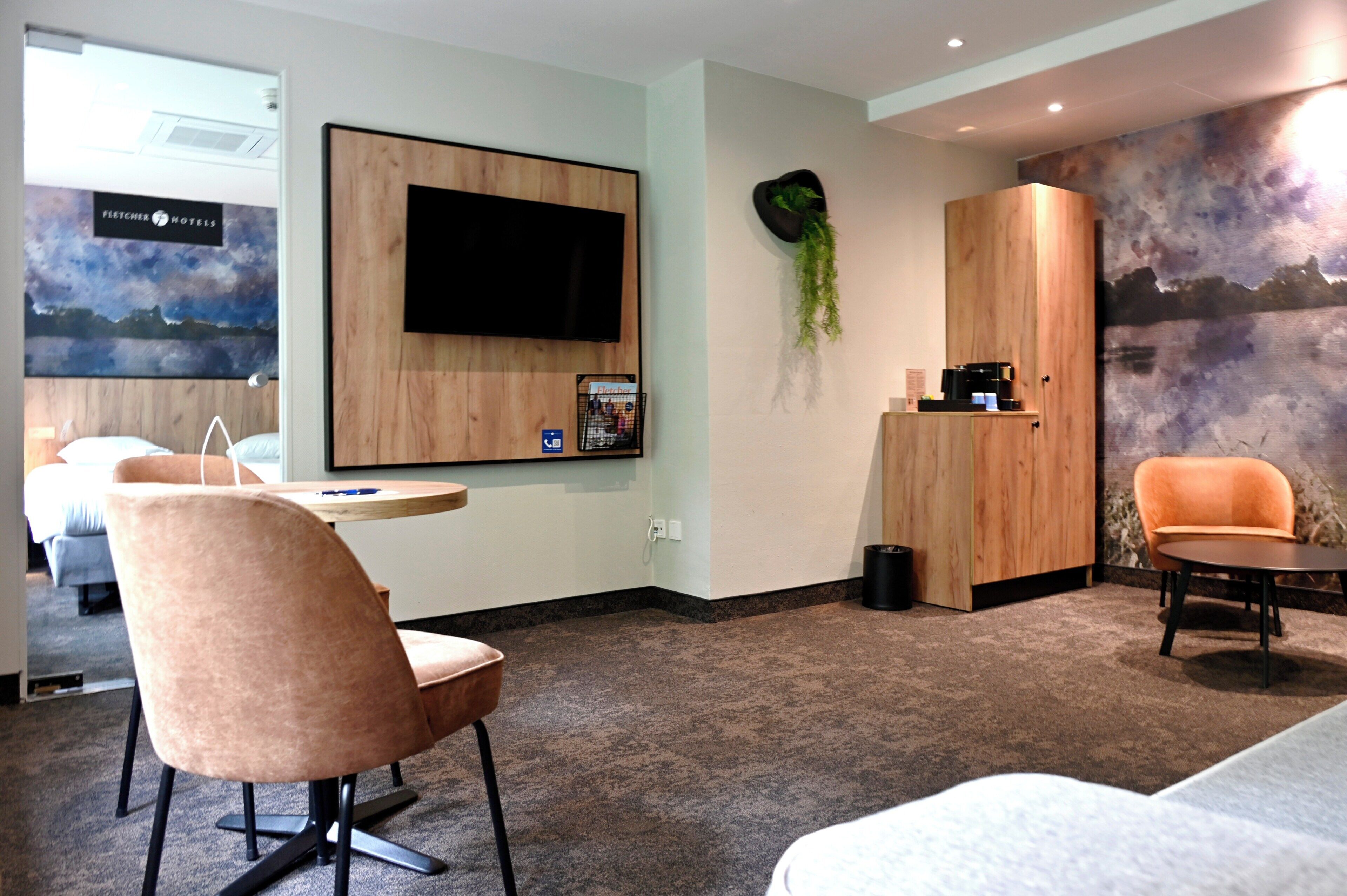 Ein Hotelzimmer mit Flachbildfernseher, Schreibtisch mit Stuhl, einer Sitzecke und einer Wandpflanze.