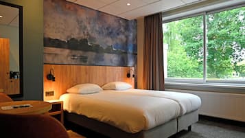 Habitación de hotel con una cama grande, un cuadro en la pared, una ventana con vistas a árboles y una mesita con una silla.