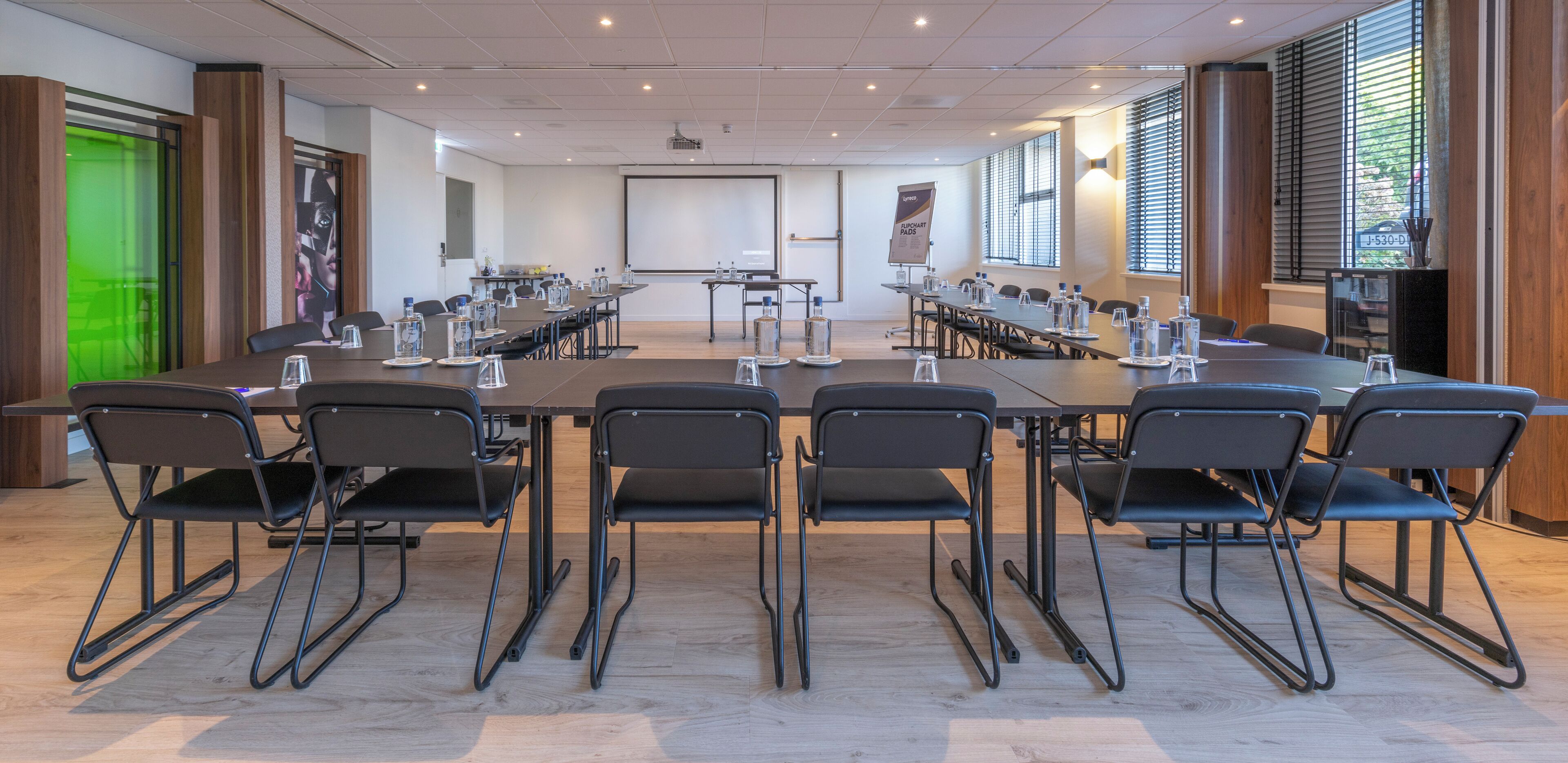 Een vergaderzaal met een grote tafel, meerdere stoelen, een projectiescherm en een presentatiegedeelte.