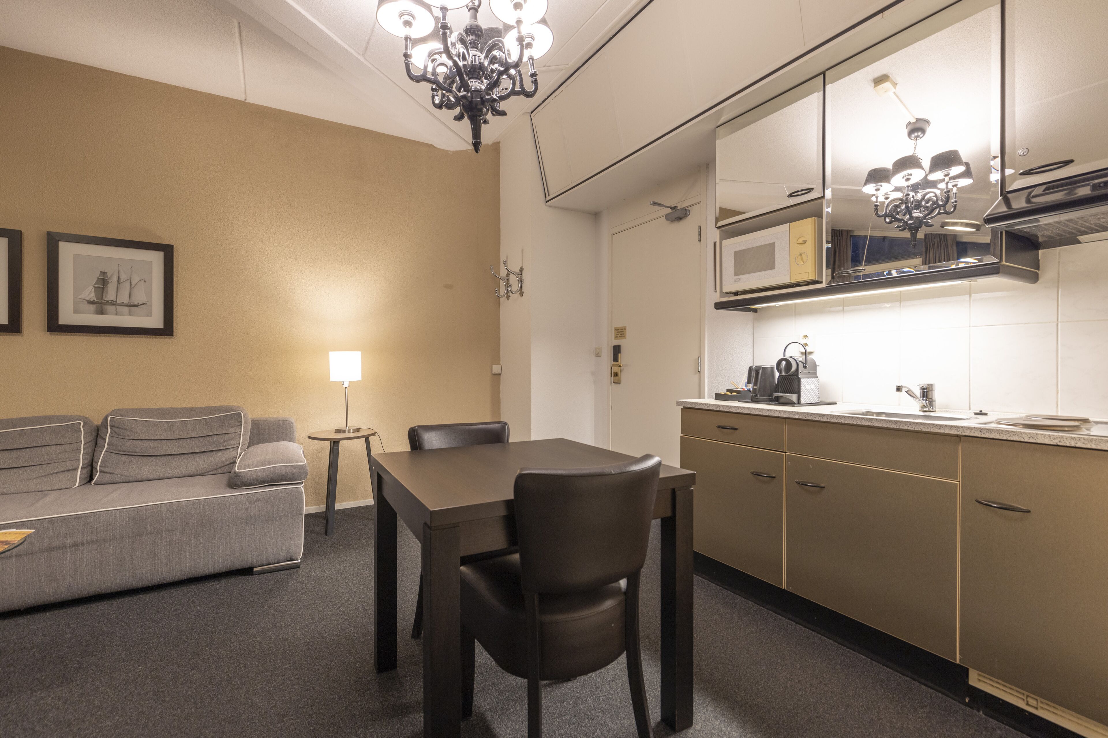 A compact hotel room with a kitchenette, a dining area with a table and chairs, a sofa, and a chandelier.