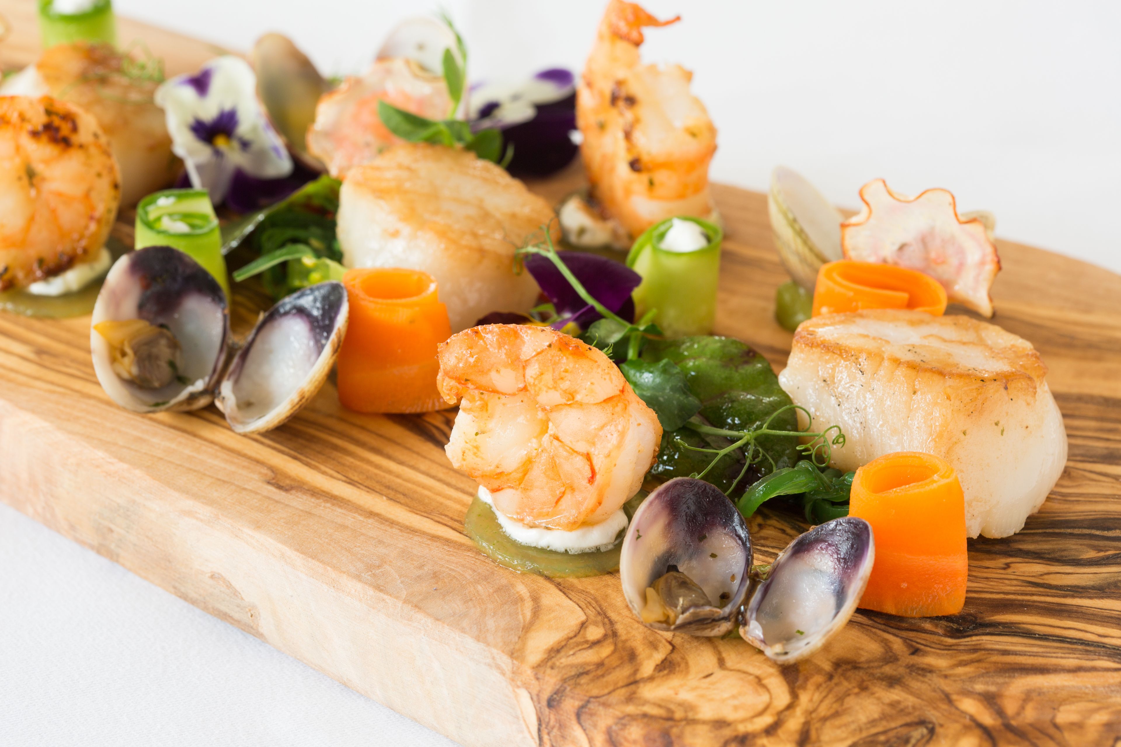 A wooden board with various seafood dishes, including shrimp, scallops, and mussels, garnished with herbs and vegetables.