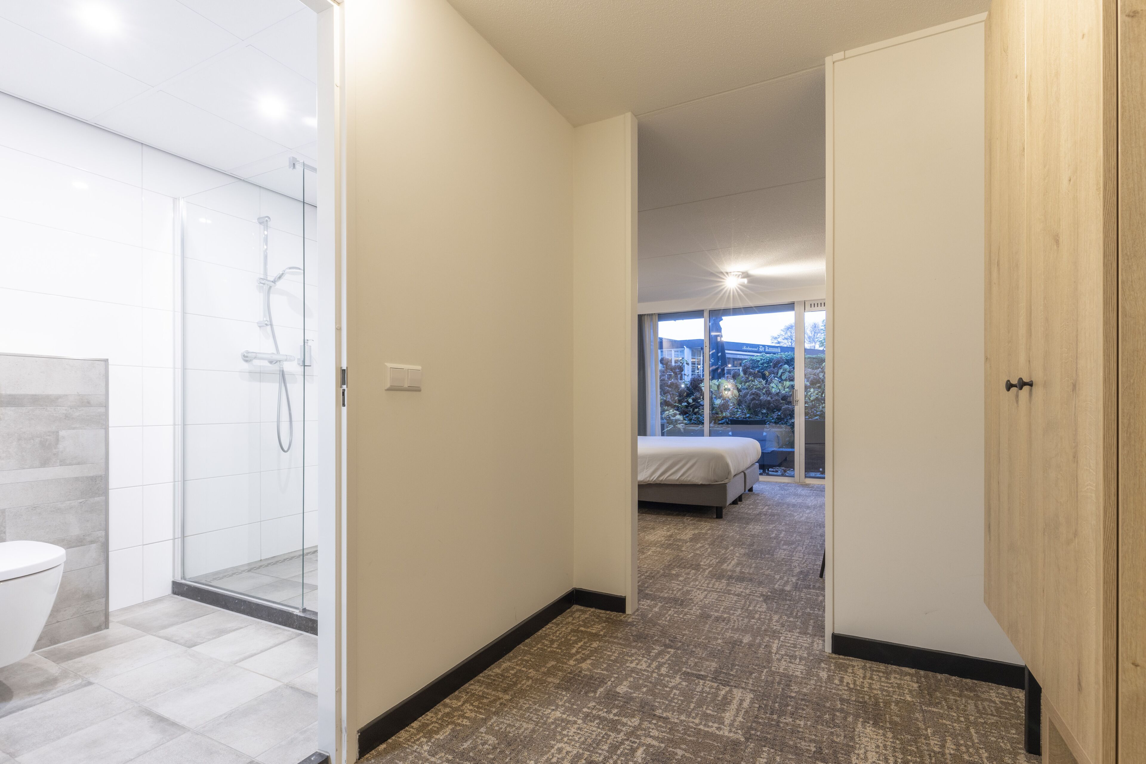 A modern hotel room with a shower, toilet, and a bed with a view of the outdoors.