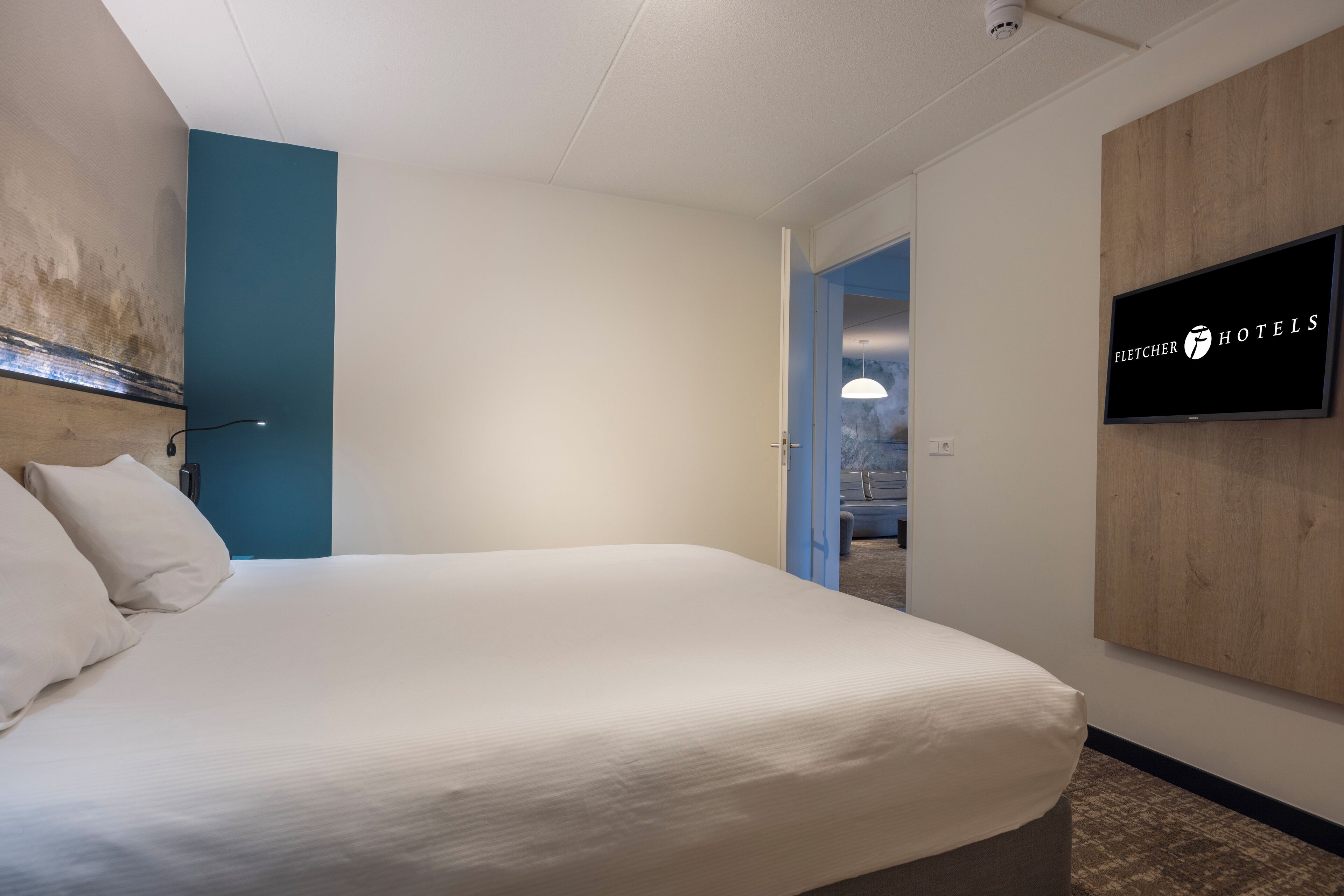 A hotel room with a large bed, a television mounted on the wall, and a view into the adjacent room.