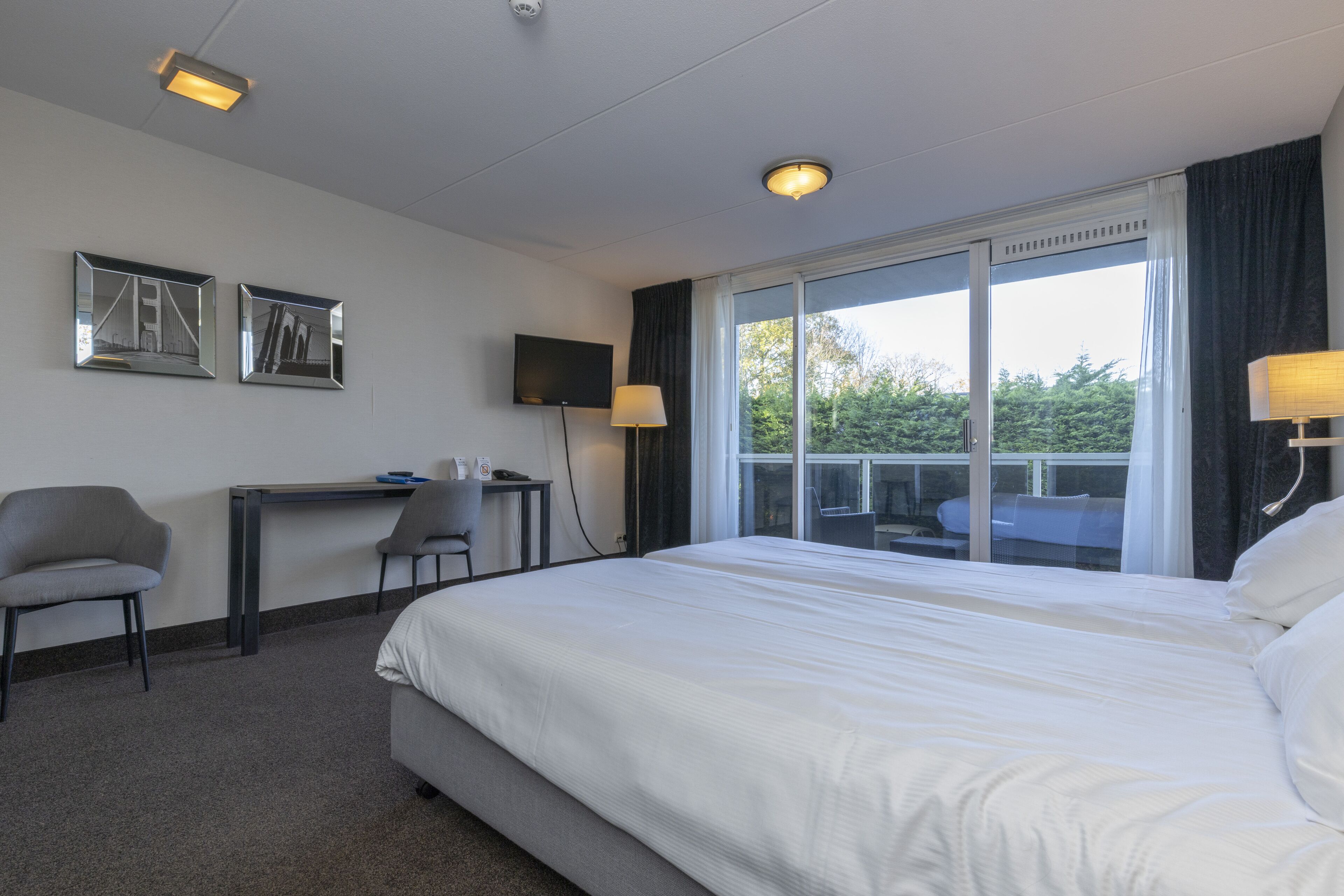 Quarto de hotel com uma cama grande, uma mesa com cadeira, uma televisão e uma janela com vista para o verde.